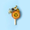 Close-up of a dried flower buttonhole with a orange bloom surrounded by blue, white and blue stems on a soft blue background.
