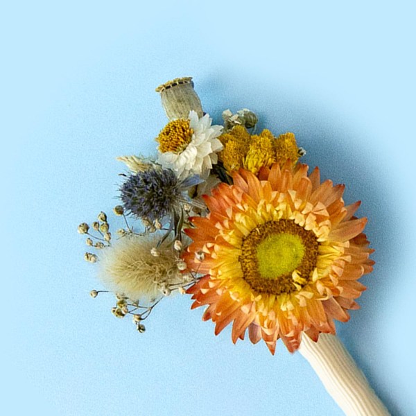 Close-up of a dried flower buttonhole with a orange bloom surrounded by blue, white and blue stems on a soft blue background.