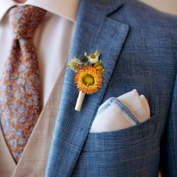 Dried flower buttonhole with a bright-orange strawflower, yellow and blue accents pinned to a blue grooms suit jacket.