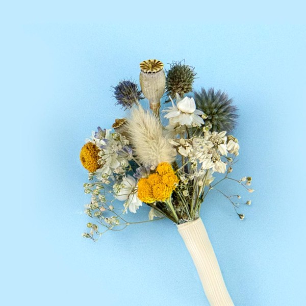 Close up of a dried flower buttonhole with yellow and blue blooms with white accents, wrapped with a white ribbon against a soft blue background